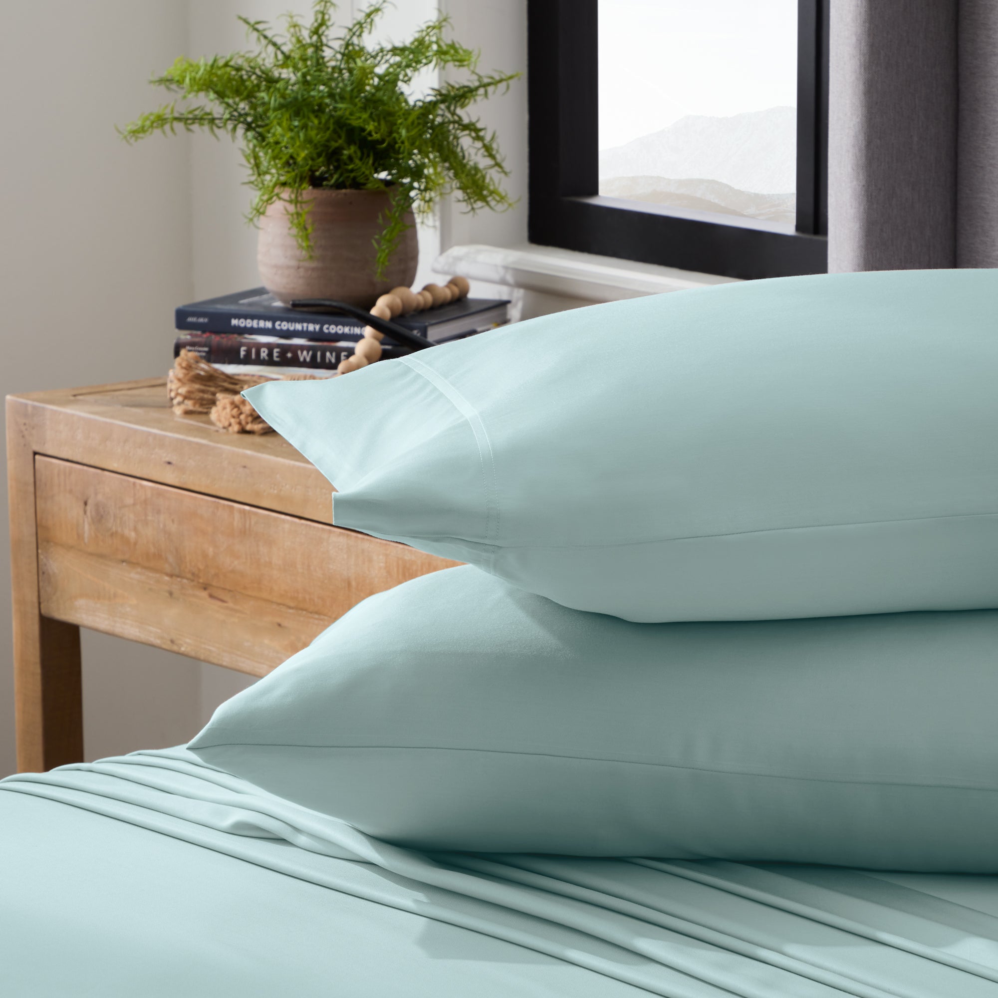 Stack of light blue pillows on a bed with a wooden nightstand and plant in the background