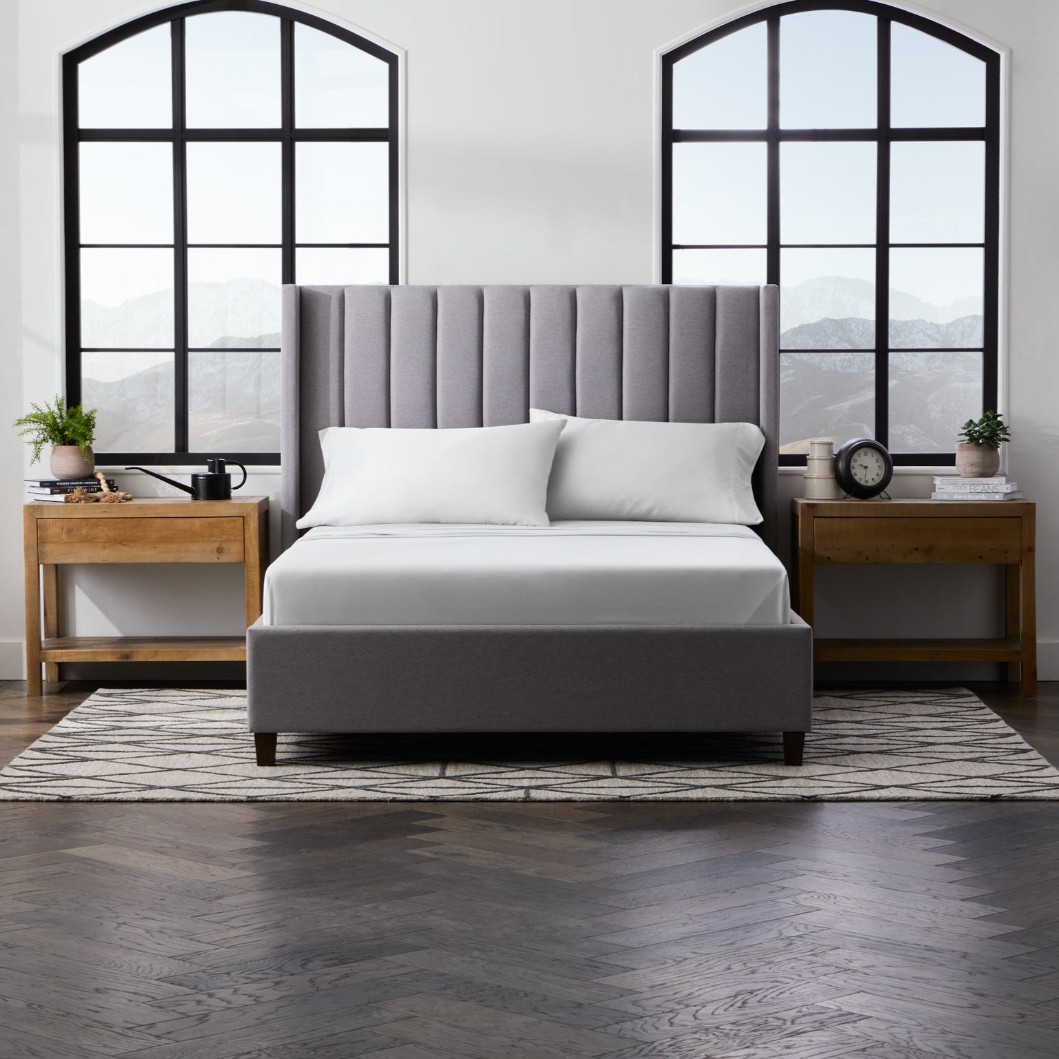 Gray upholstered bed with white bedding in a room with large windows